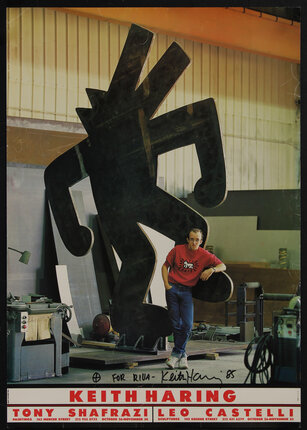 a young man with glasses in a warehouse with equipment leaning on a sculpture of an abstract flat human figure.